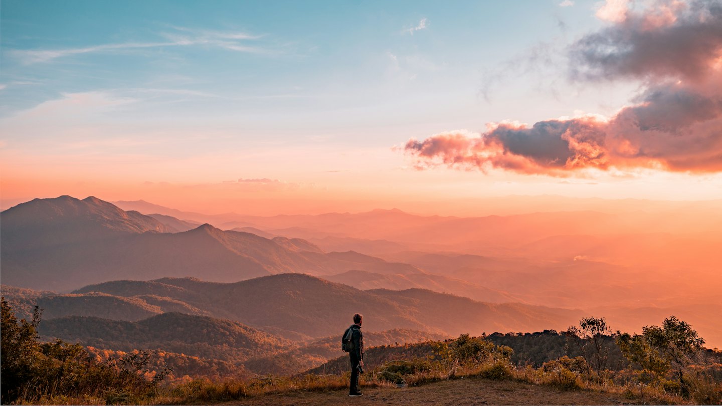 Hiker on mountain ridge overlooking hazy range at sunset.