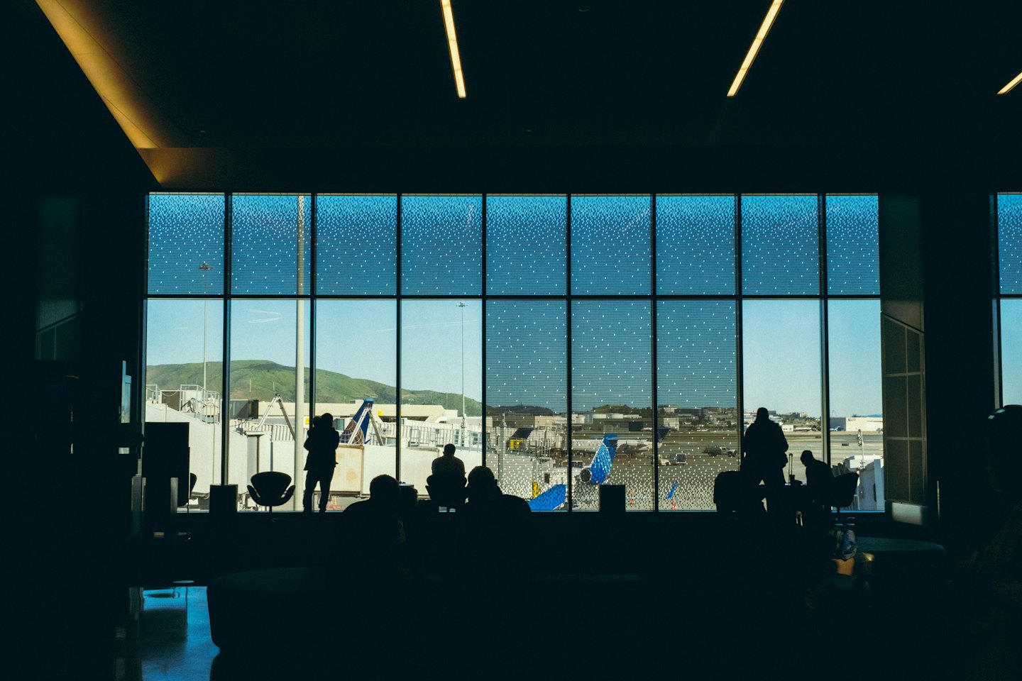 Travelers watch airplanes on the tarmac from an airport lounge.
