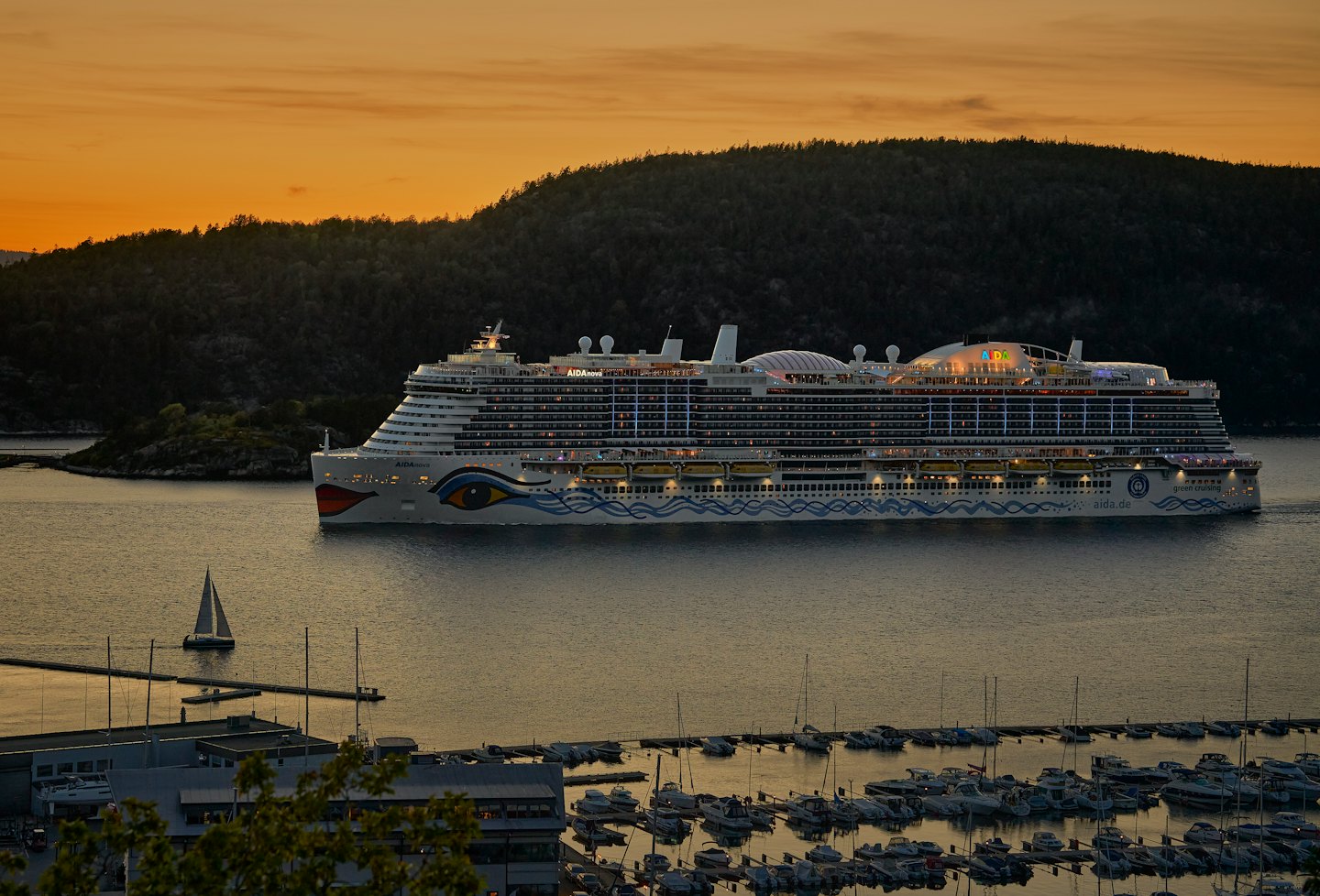 A decorated cruise ship sails past a marina at sunset.